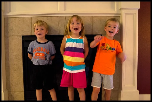 Three smiling children standing together in front of a fireplace